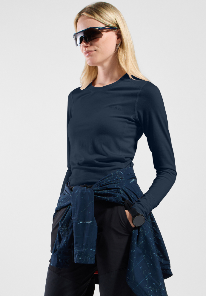 Blonde woman in a navy long-sleeve base layer, black trousers, sunglasses, and a patterned jacket tied at her waist.