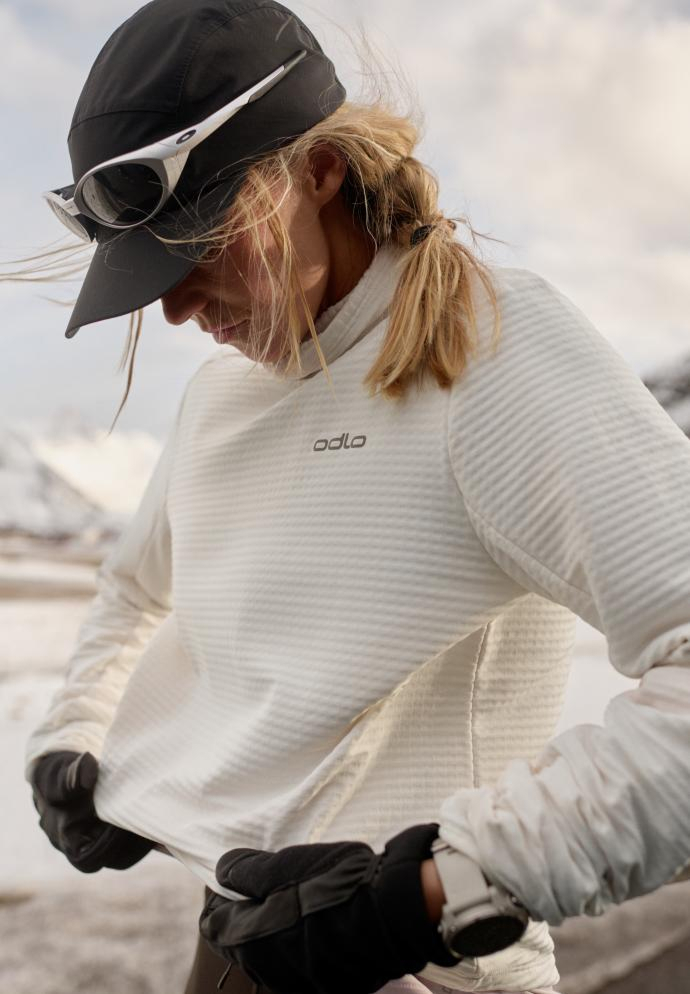 Blonde woman in snow adjusting her white Odlo mid layer. She wears a black cap, sunglasses on her head, and black gloves.
