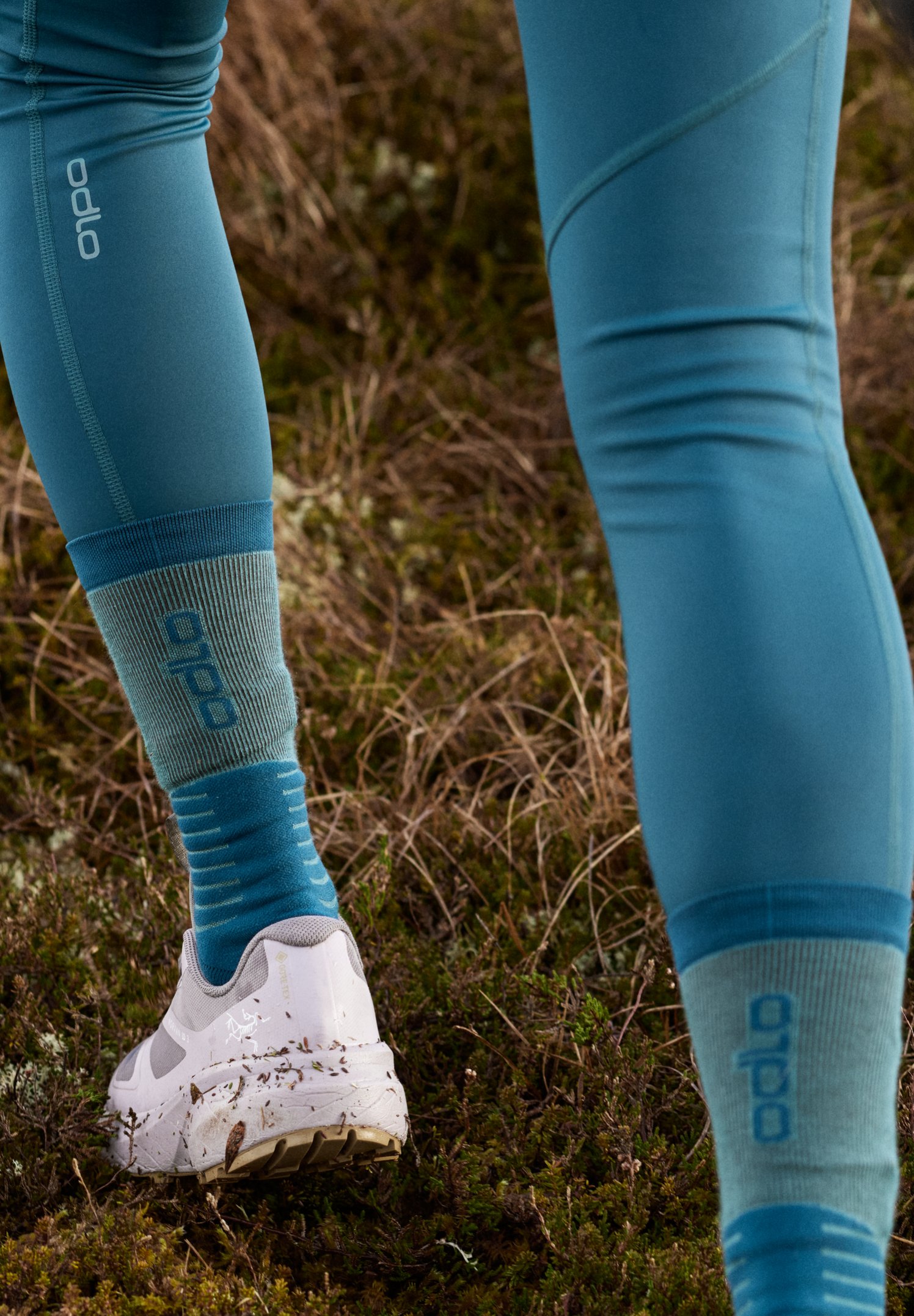 Personne enfilant des chaussettes de compression roses et violettes montant jusqu'aux genoux, par-dessus un legging clair, sur fond blanc.