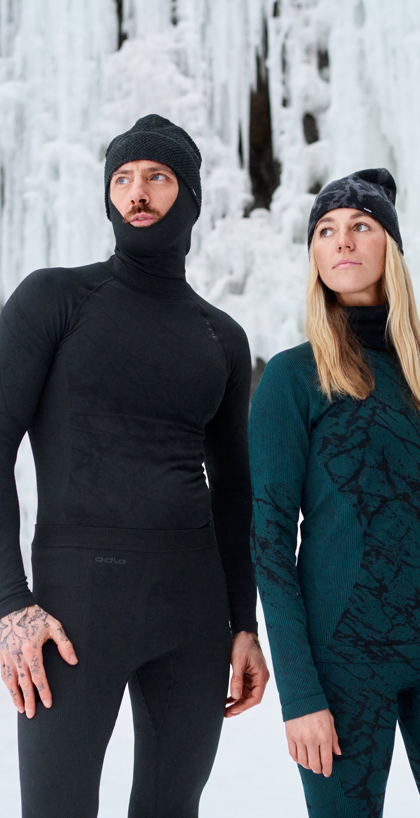 Two people in winter base layers and beanies stand in snowy terrain before an ice-covered cliff, looking off-camera in cold, overcast light.