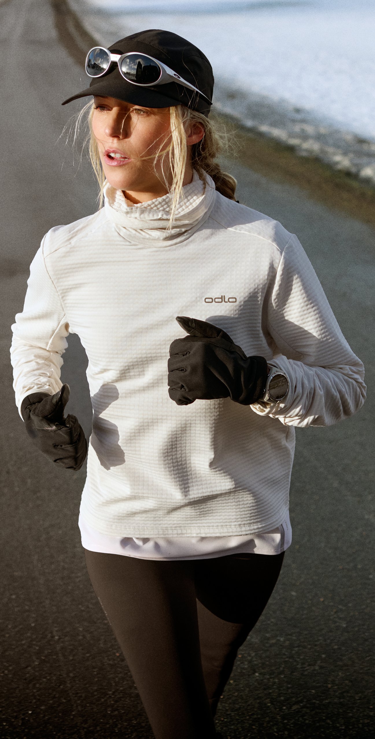 Vrouw rent op een verharde weg met sneeuw langs de kant. Ze draagt een wit Odlo-shirt, een zwarte pet, een zonnebril en zwarte handschoenen.