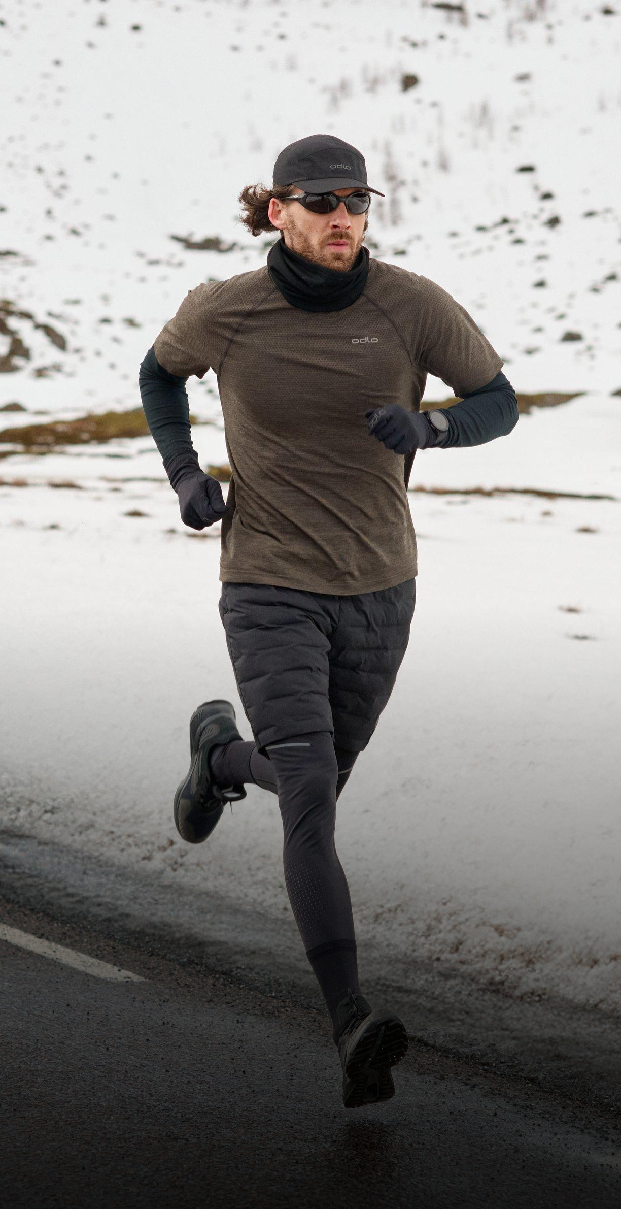Hardloper in winterkleding en met zonnebril joggend op een natte weg langs besneeuwde velden en heuvels onder een bewolkte lucht.
