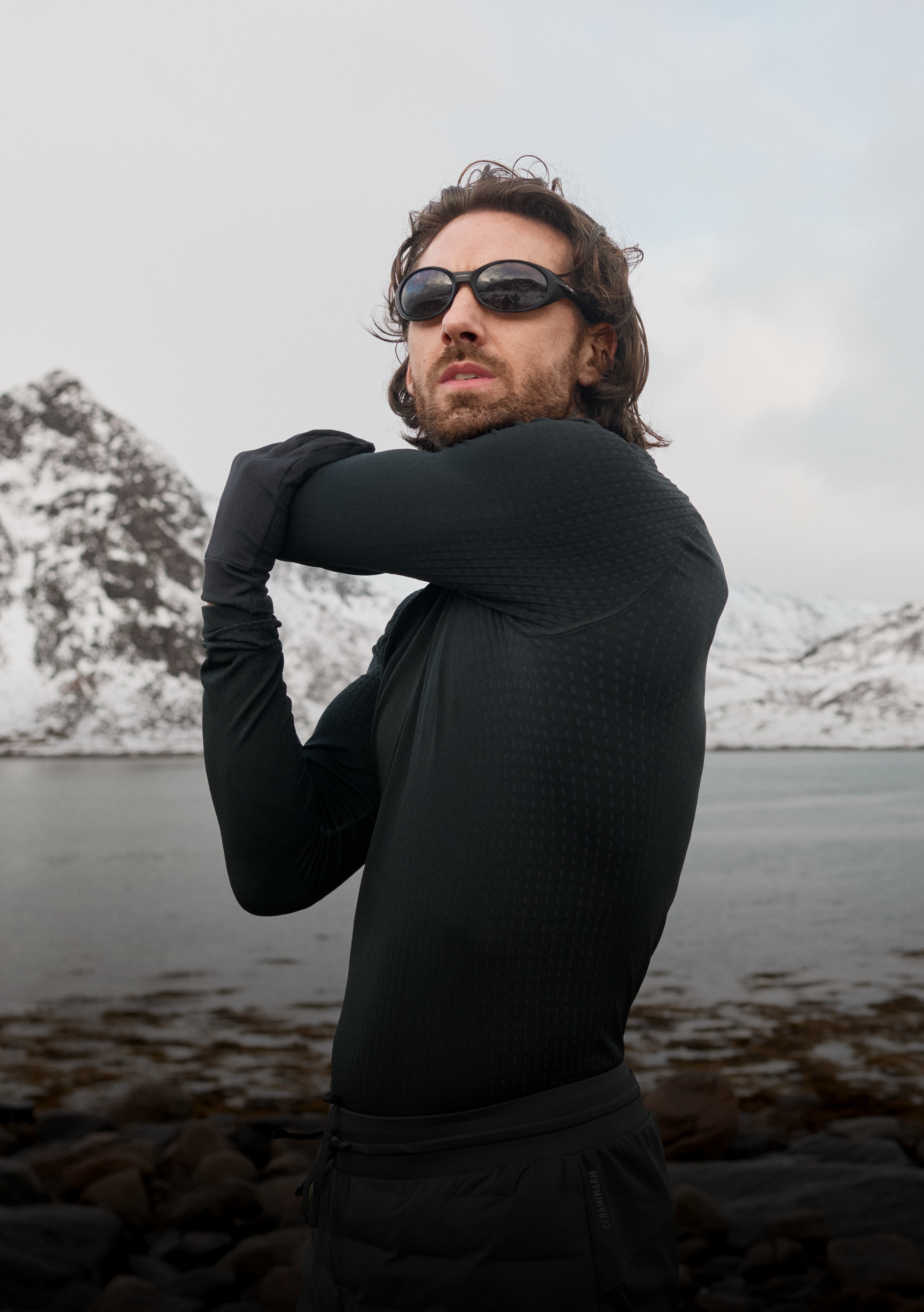 Bärtiger Mann in schwarzem Performance Warm Base Layer und Sonnenbrille streckt seinen Arm an einem felsigen Ufer vor einer Kulisse schneebedeckter Berge aus.