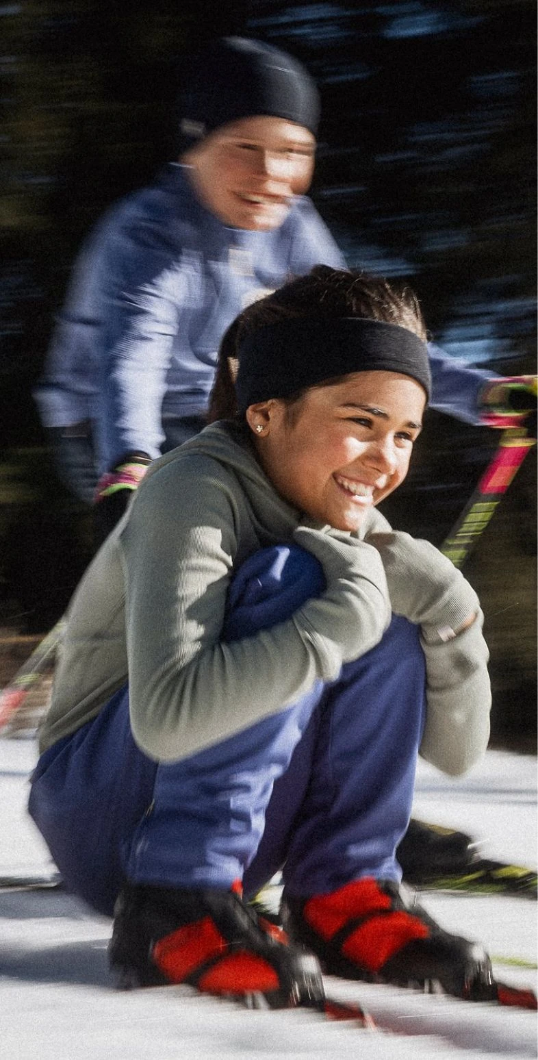 Lächelndes Mädchen in grauem Base Layer, blauer Hose und roten Stiefeln kauert im Schnee. Im Hintergrund ist unscharf ein vorbeifahrender Skifahrer zu sehen.