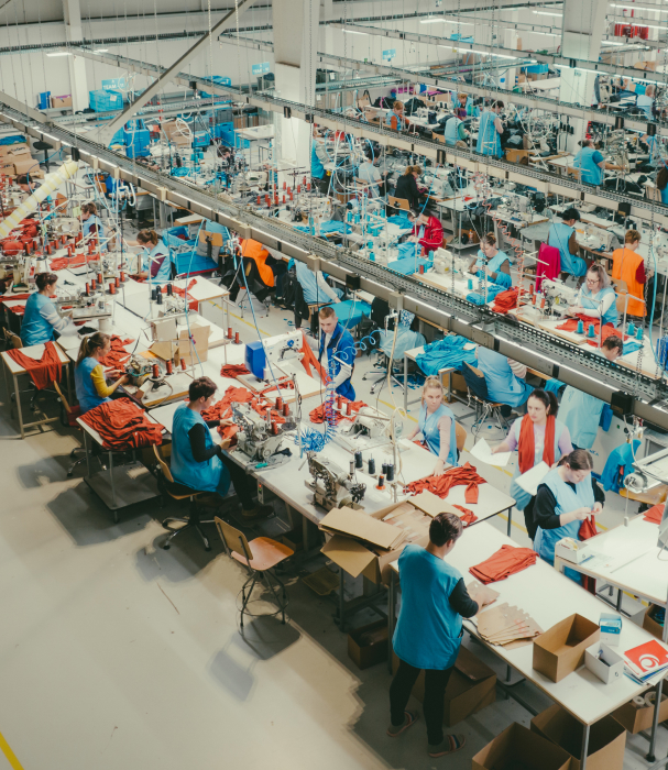 Blick von oben auf eine große Bekleidungsfabrik. Viele Arbeiter in blauen Uniformen sitzen an langen Tischen und nähen roten Stoff.