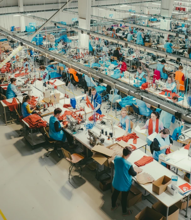 Blick von oben auf eine große Bekleidungsfabrik. Viele Arbeiter in blauen Uniformen sitzen an langen Tischen und nähen roten Stoff.