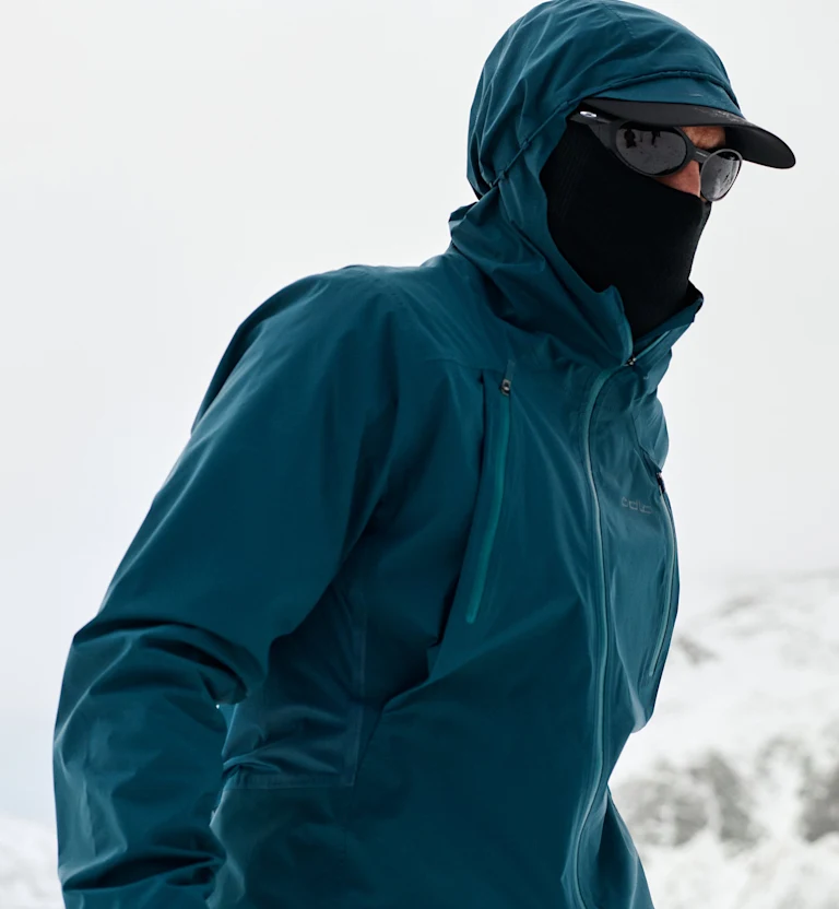 Person in einer dunkeltürkisfarbenen Kapuzenjacke, mit schwarzer Gesichtsmaske, Cap und Sonnenbrille in einer verschneiten Landschaft.