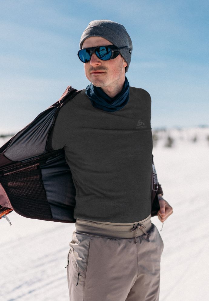 Man in a snowy landscape wearing a beanie, blue sunglasses, dark grey top and beige trousers, holding his jacket open.
