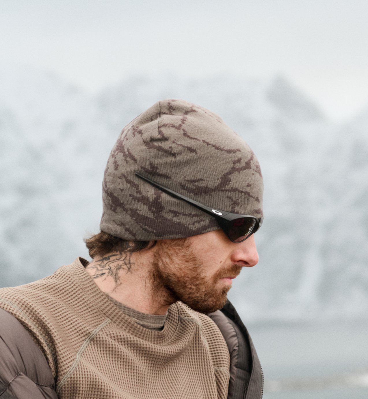 Profilo di un uomo con la barba e un tatuaggio sul collo, berretto mimetico marrone, occhiali da sole e maglietta color cammello, con montagne innevate sullo sfondo.