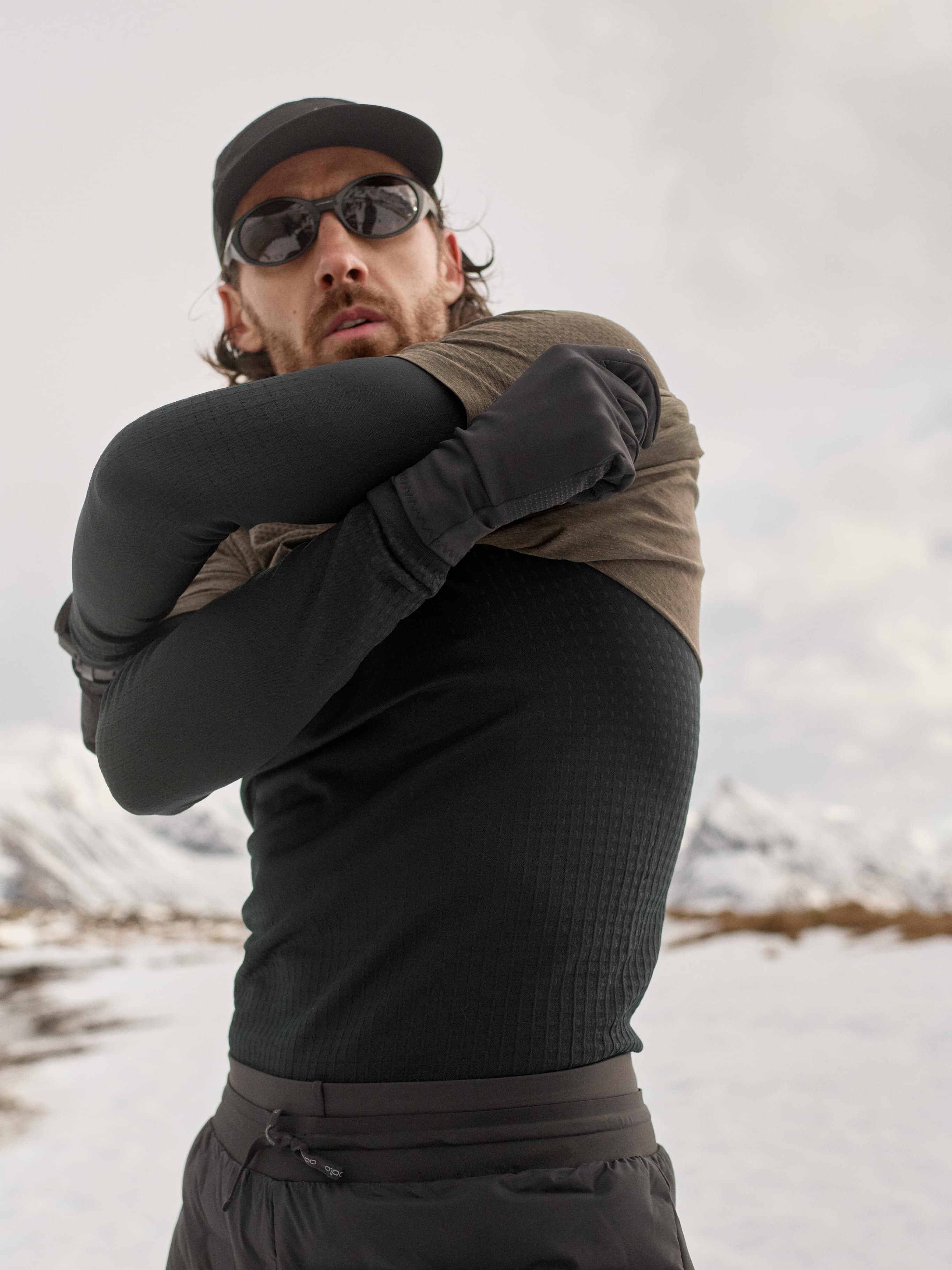 Bearded man in cap and sunglasses pulling an dark brown running t-shirt over a black performance warm base layer in a snowy mountain setting.
