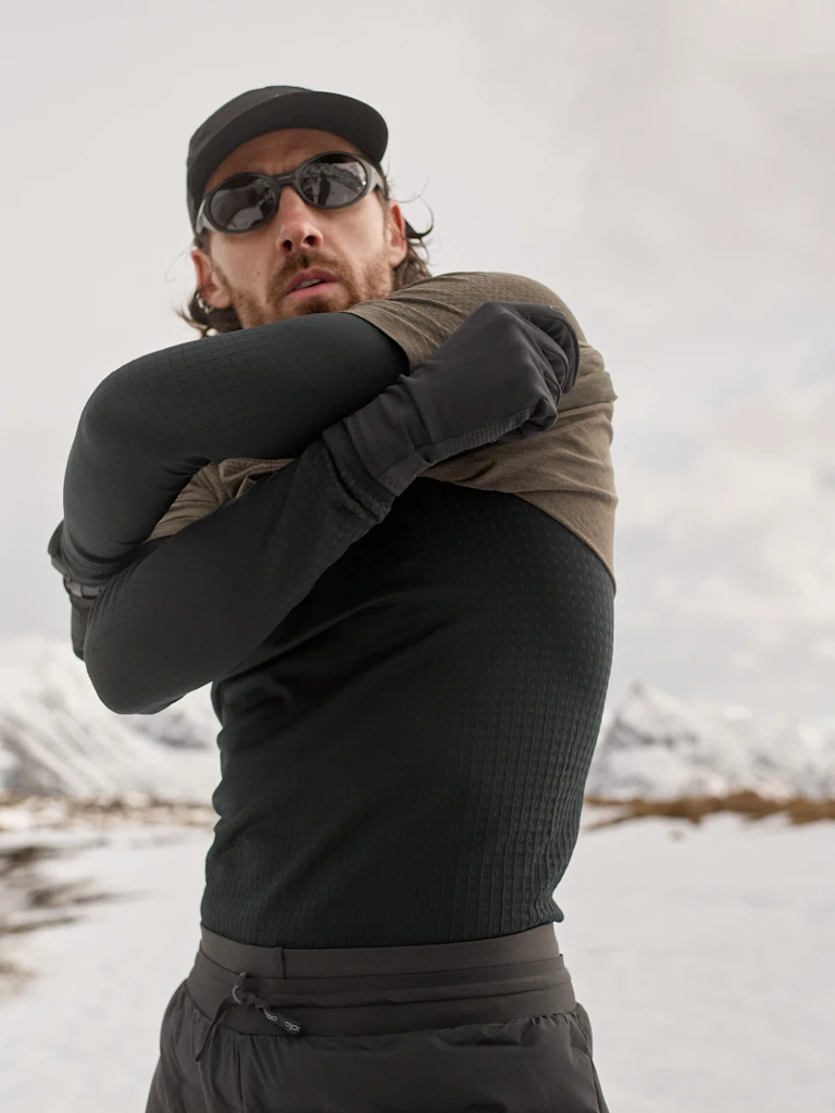 Bearded man in cap and sunglasses pulling an dark brown running t-shirt over a black performance warm base layer in a snowy mountain setting.