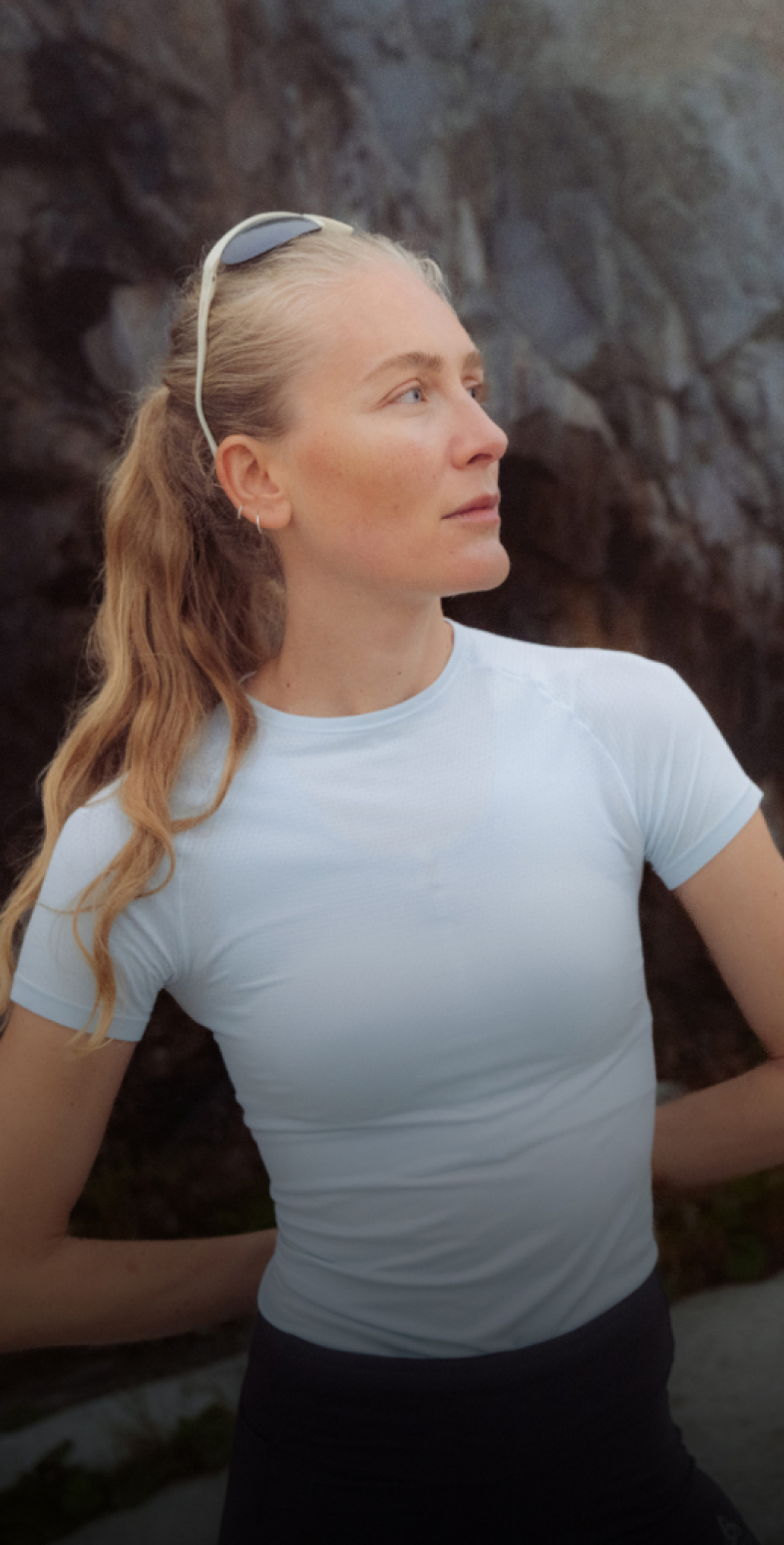 A blonde woman in a light blue base layer with sunglasses on her head looks to the side against a dark rocky background.