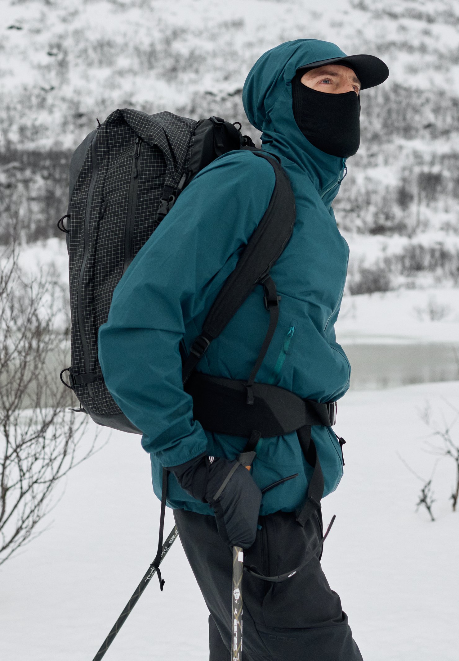 Wanderer in tealfarbener Jacke, schwarzer Gesichtsmaske und großem Rucksack hält Trekkingstöcke in einer verschneiten Winterlandschaft.