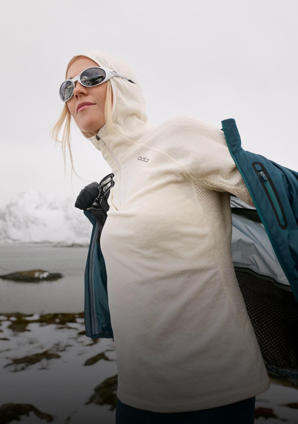 Frau mit schwarzem Cap, Sonnenbrille, Handschuhen und weißem Odlo-Top richtet ihr Shirt in einer verschneiten Landschaft.