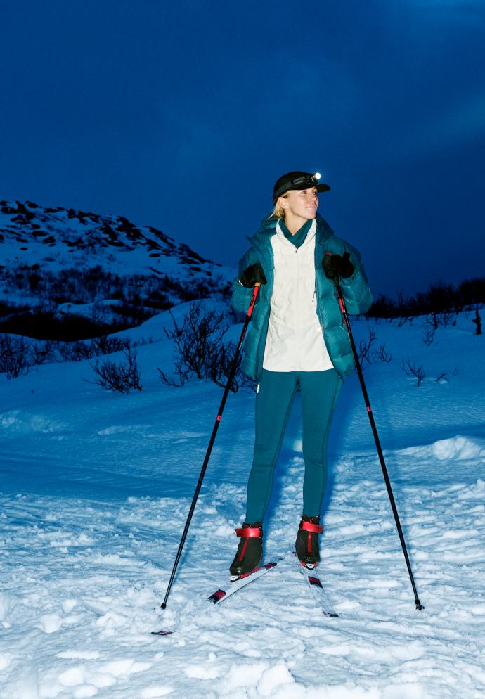 Femme faisant du ski de fond de nuit dans un paysage enneigé, portant une veste bleue, un legging foncé et une lampe frontale allumée.