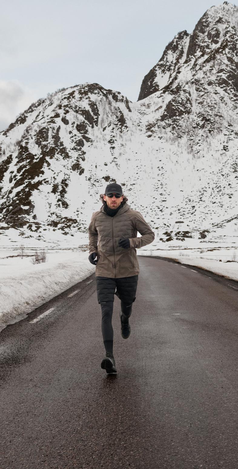 Un homme en veste marron et legging noir court sur une route mouillée vers l'appareil photo, avec des montagnes enneigées en arrière-plan.