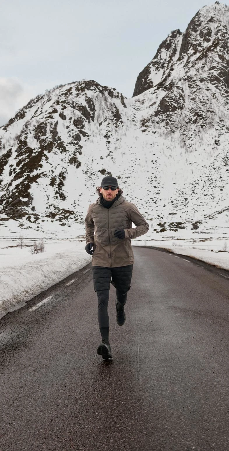 Un homme en veste marron et legging noir court sur une route mouillée vers l'appareil photo, avec des montagnes enneigées en arrière-plan.