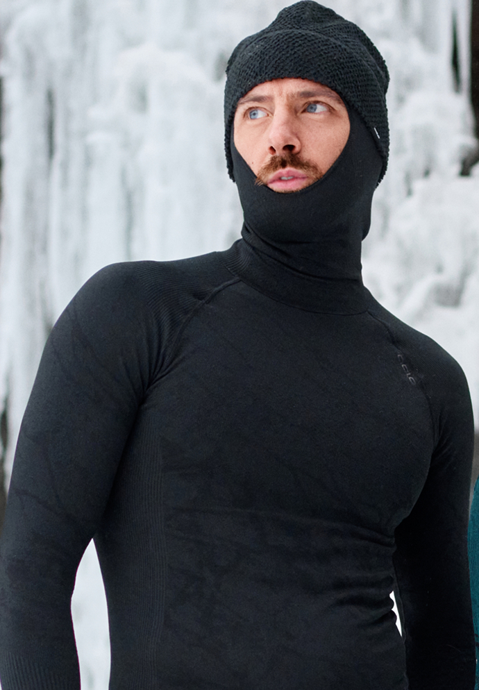 Man in a black Odlo thermal base layer top with a balaclava and beanie, looking aside against a frozen, icy background.
