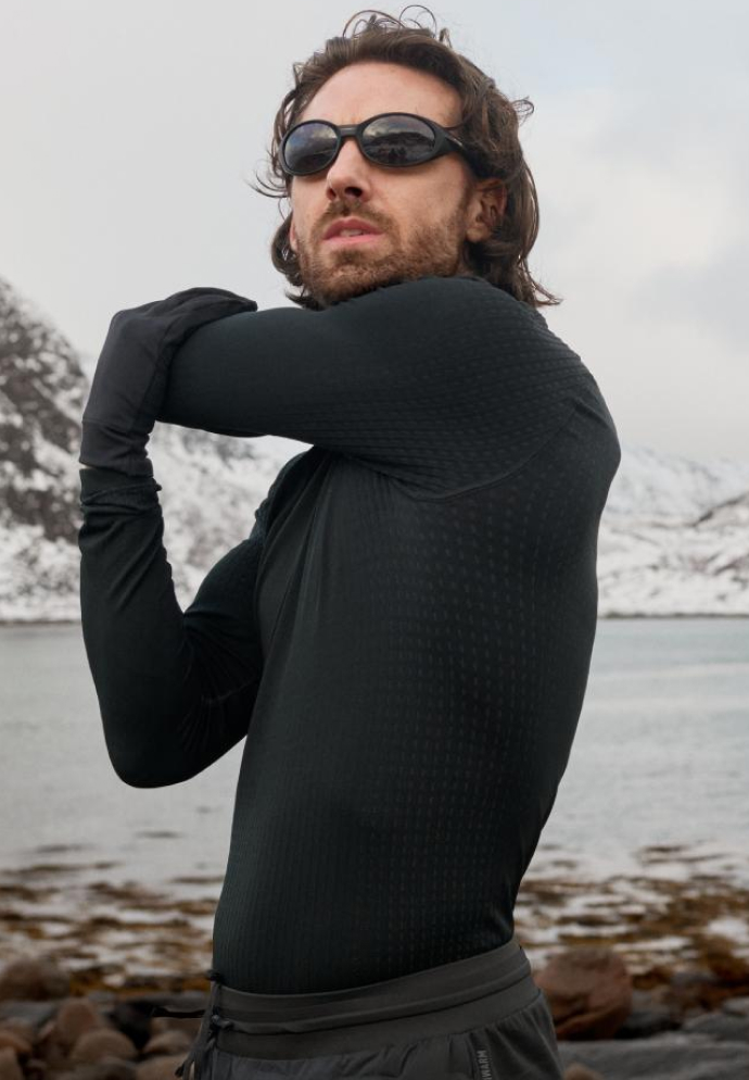 Ein Mann in schwarzem sportlichem Base Layer und Sonnenbrille macht Dehnübungen an einem felsigen Ufer mit Wasser und schneebedeckten Bergen im Hintergrund.