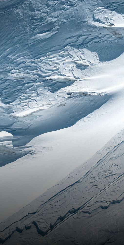 Luftaufnahme von zwei Skifahrern, die einen steilen, strukturierten weißen Schneegrat hinaufsteigen und dabei lange, dunkle Schatten auf den Berg werfen.