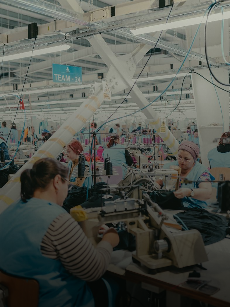 Many women in light blue vests operate sewing machines in a large, bright garment factory, working on dark fabric.