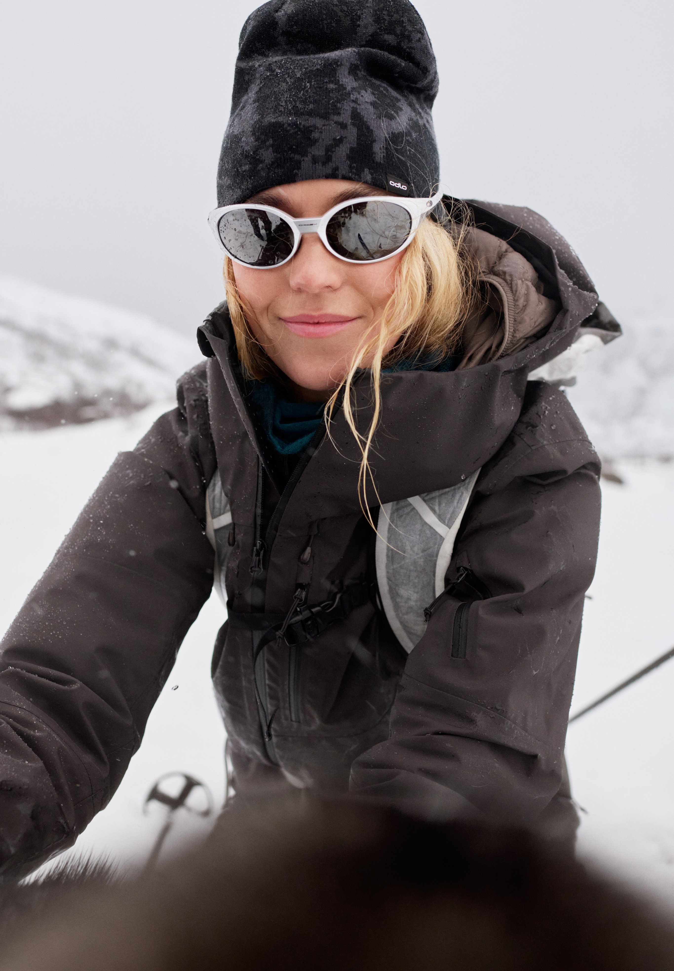 Lachende blonde vrouw die een selfie maakt in de sneeuw. Ze draagt een zwarte muts, een witte zonnebril en een donkere winterjas.