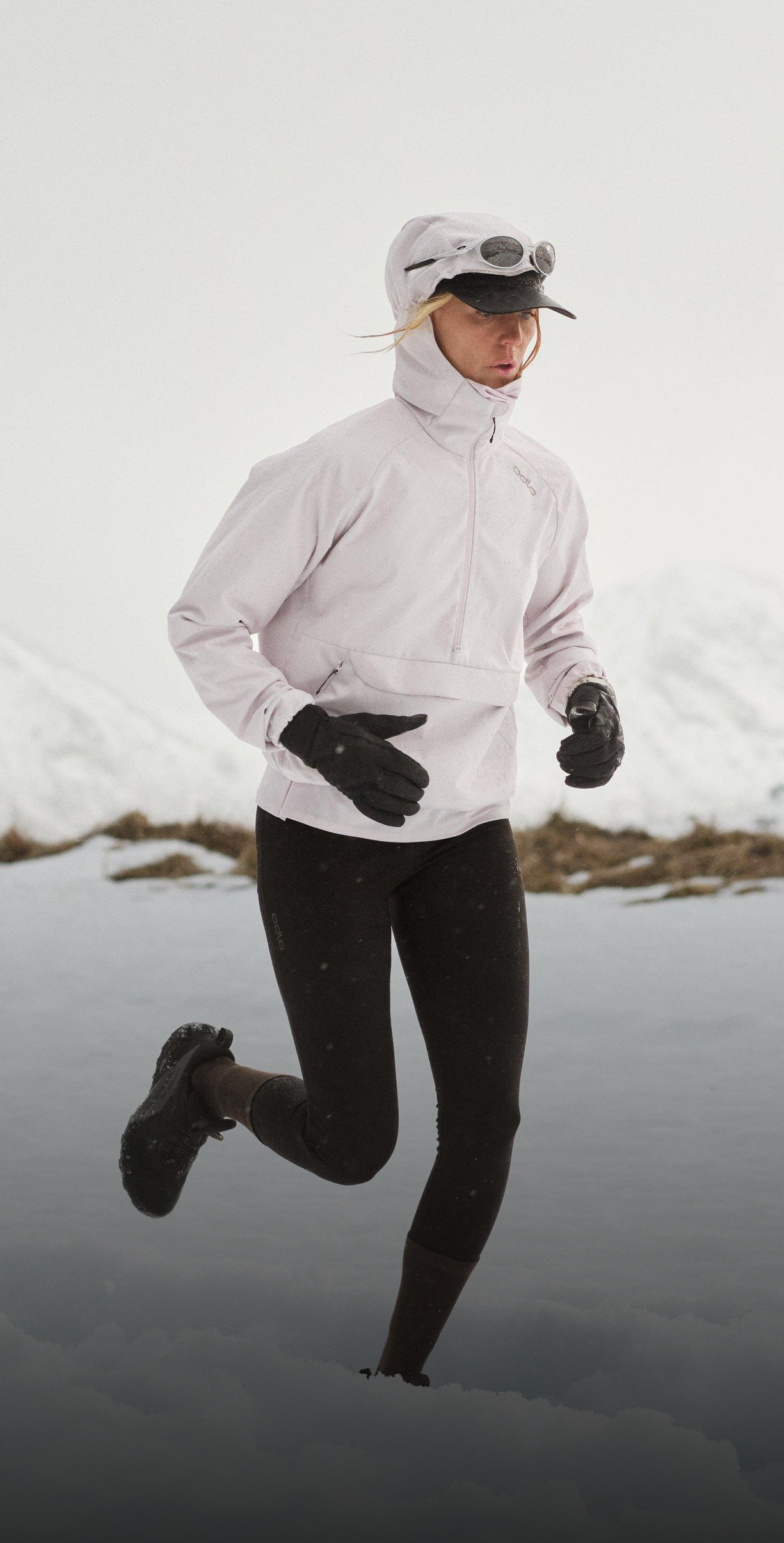 Runner in pale jacket, black leggings, and gloves jogging through snowy terrain, wearing a hood, cap, and goggles, with mountains behind.