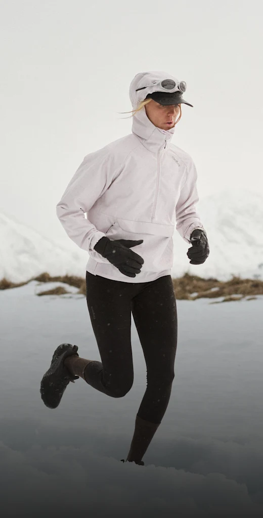 Runner in pale jacket, black leggings, and gloves jogging through snowy terrain, wearing a hood, cap, and goggles, with mountains behind.