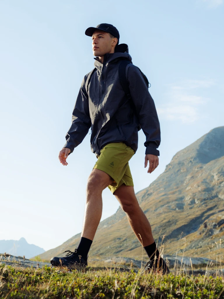 Mann beim Wandern in den Bergen, der eine dunkle Jacke, grüne Shorts, eine schwarze Kappe und Schuhe unter einem blauen Himmel trägt.