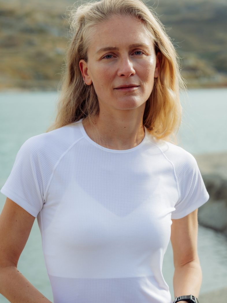 Blonde woman in a white light base layer looking at the camera, with a blurred lake and rocky hills behind her.