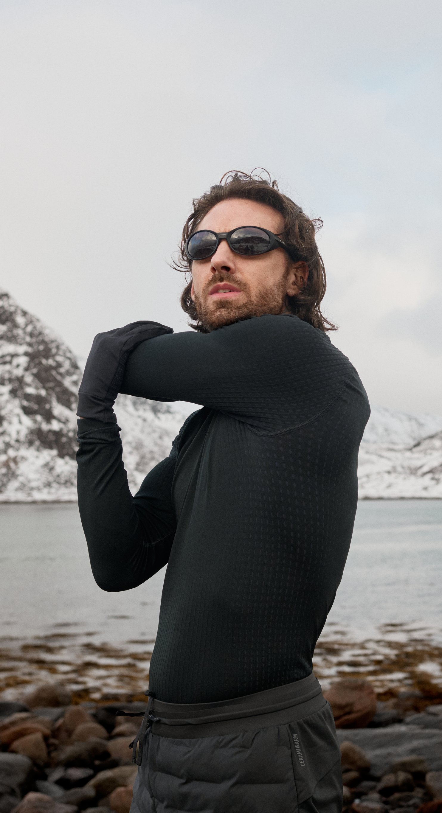 Mann in schwarzem, wärmendem Performance Base Layer und mit Sonnenbrille streckt auf einem felsigen Ufer den Arm aus, hinter ihm schneebedeckte Berge und Wasser.