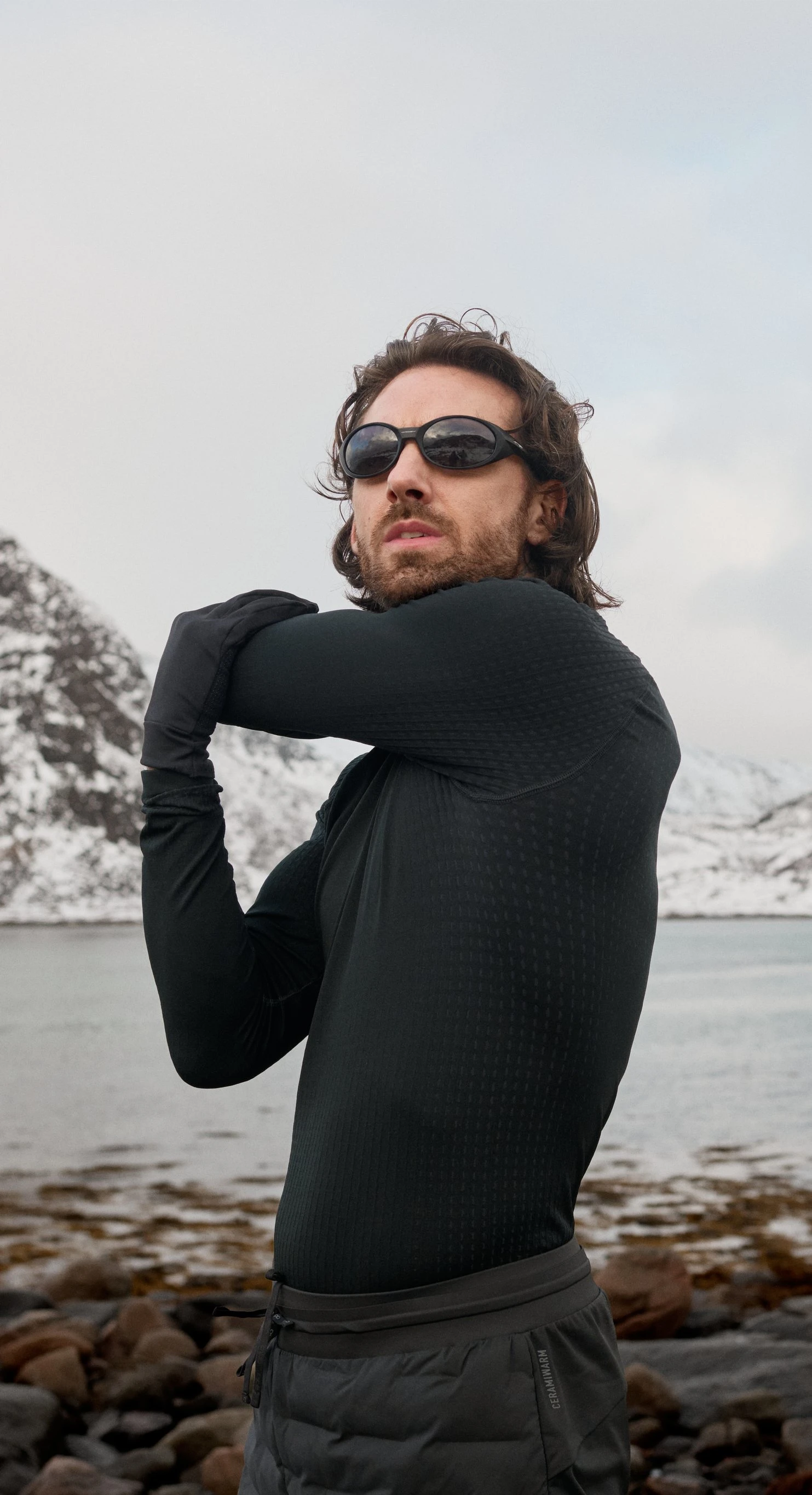 Mann in schwarzem, wärmendem Performance Base Layer und mit Sonnenbrille streckt auf einem felsigen Ufer den Arm aus, hinter ihm schneebedeckte Berge und Wasser.