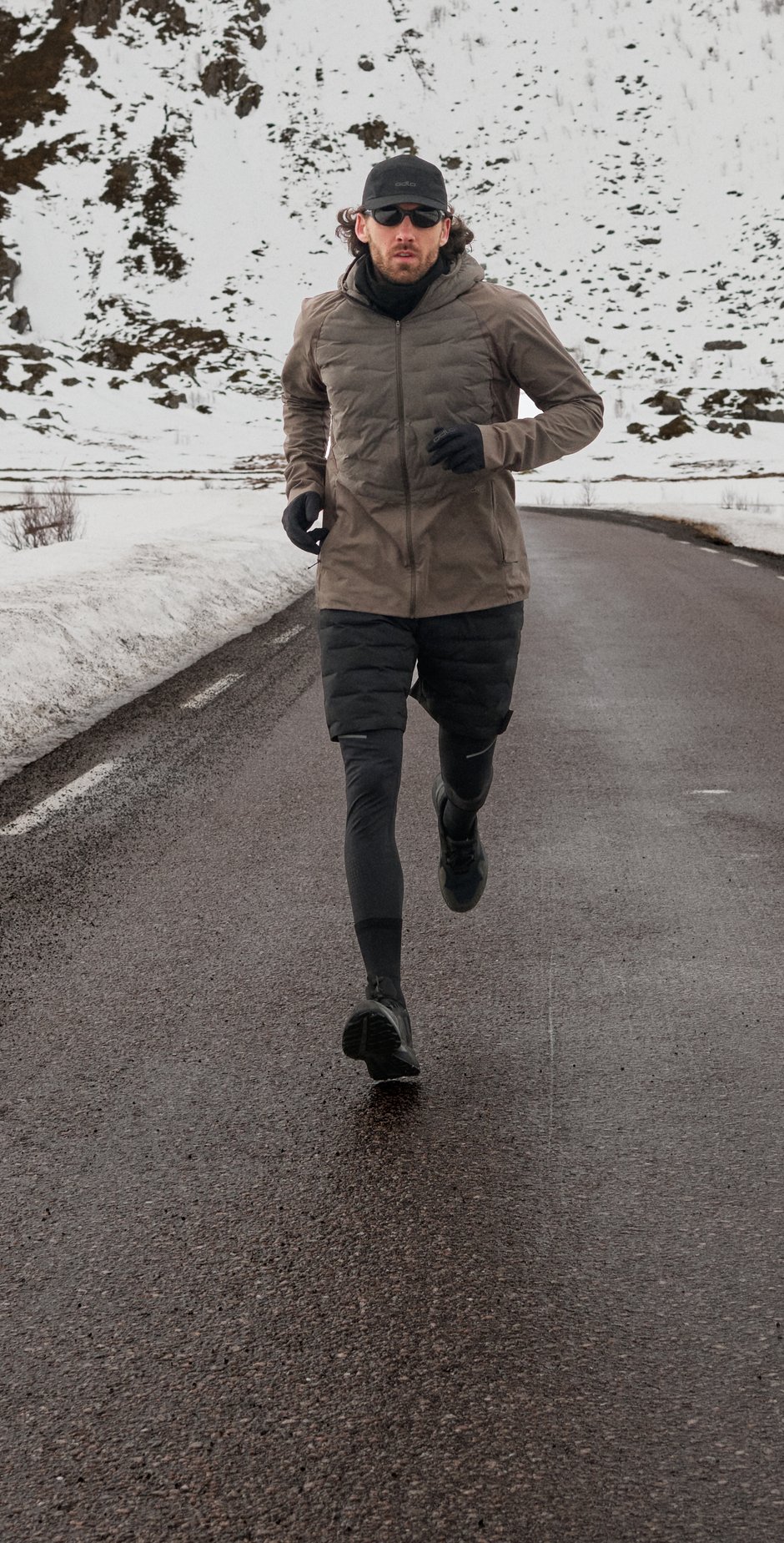 A man in a brown jacket and cap runs towards the camera on an empty road surrounded by snowy mountains.