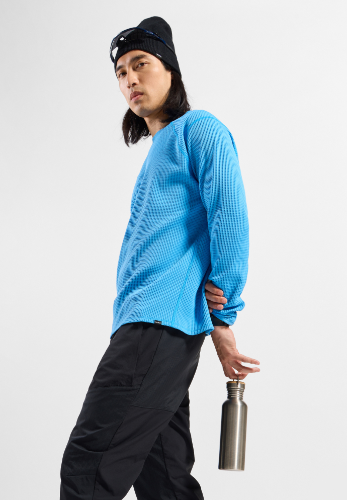Man looking back, wearing a blue mid layer, black trousers, boots and beanie, holding a metal water bottle.