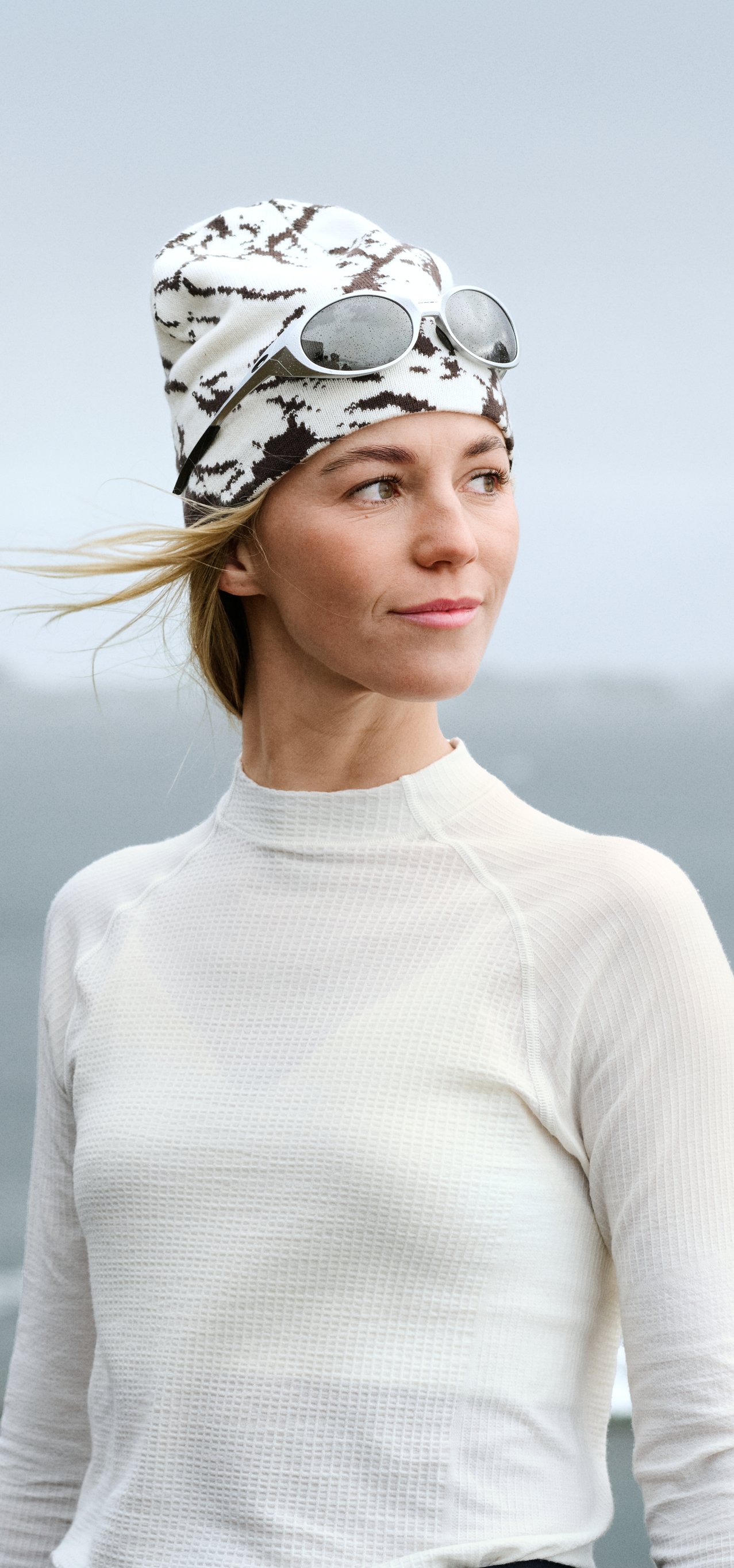 Femme en haut thermique blanc et bonnet à motifs avec lunettes de soleil regardant vers la droite. Personne floue au premier plan, mer en arrière-plan.