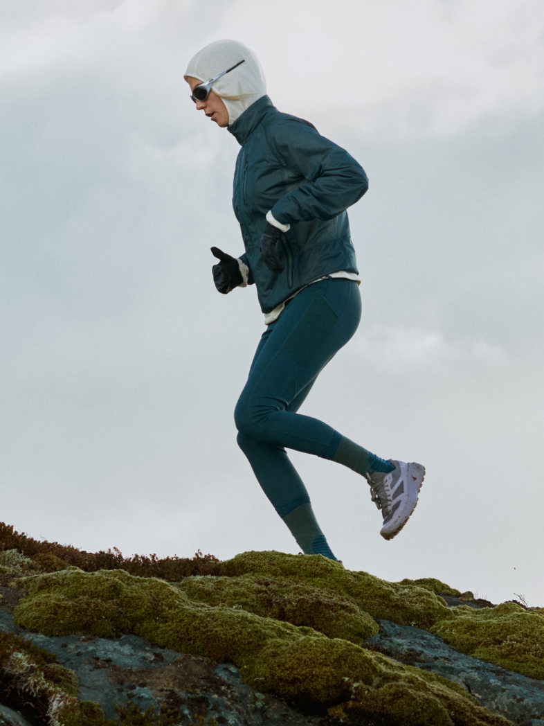 Een hardloper in een blauwgroen jack, legging en witte bivakmuts jogt over bemoste rotsen onder een grijze lucht.