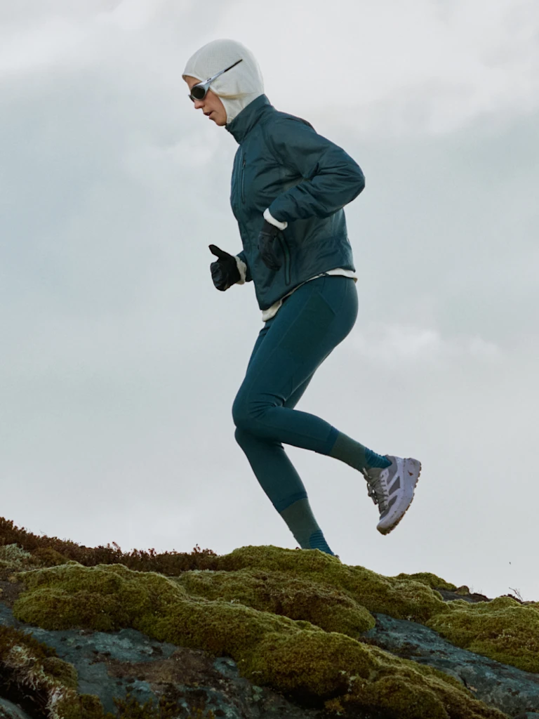 Een hardloper in een blauwgroen jack, legging en witte bivakmuts jogt over bemoste rotsen onder een grijze lucht.