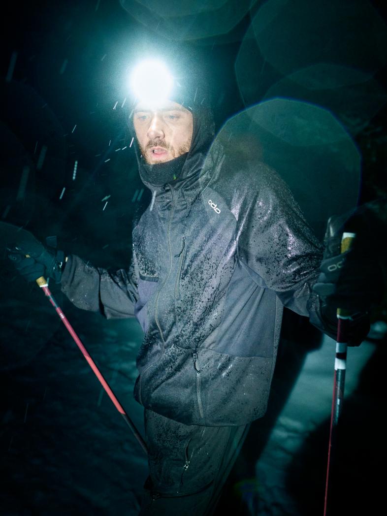 Un homme portant une veste Dual Dry noire mouillée et une lampe frontale lumineuse tient des bâtons de ski rouges la nuit, sous la neige qui tombe.