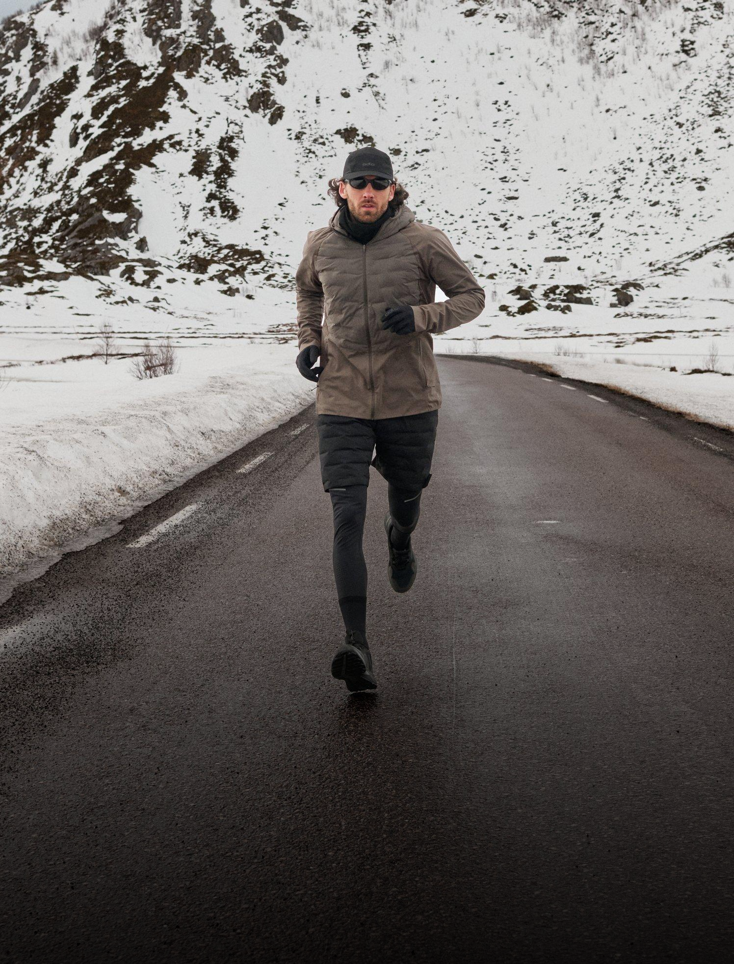Ein Mann in einer braunen Odlo Zeroweight Insolator Jacke und Shorts sowie einer Mütze läuft auf einer Straße auf die Kamera zu, mit großen schneebedeckten Bergen im Hintergrund.