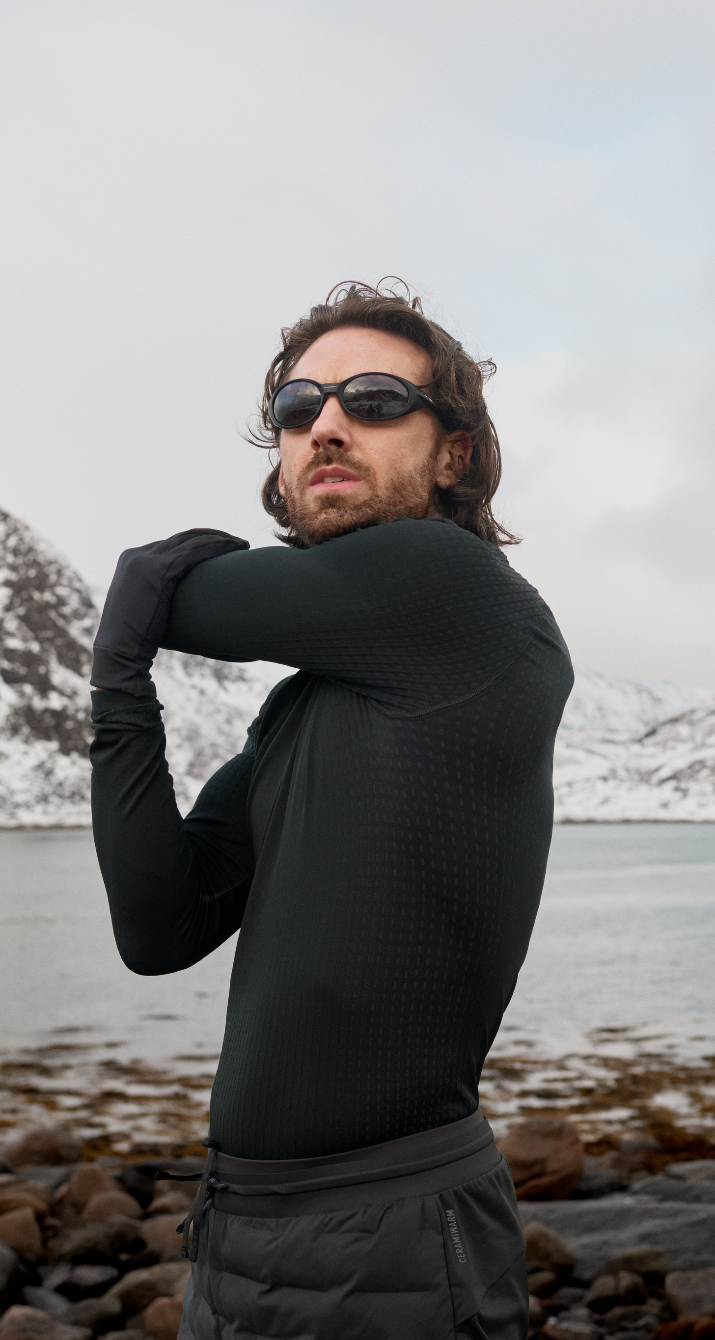 Un uomo con la barba, in abbigliamento da performance nero e occhiali da sole, allunga il braccio in uno stretching su una riva rocciosa, con montagne innevate sullo sfondo.