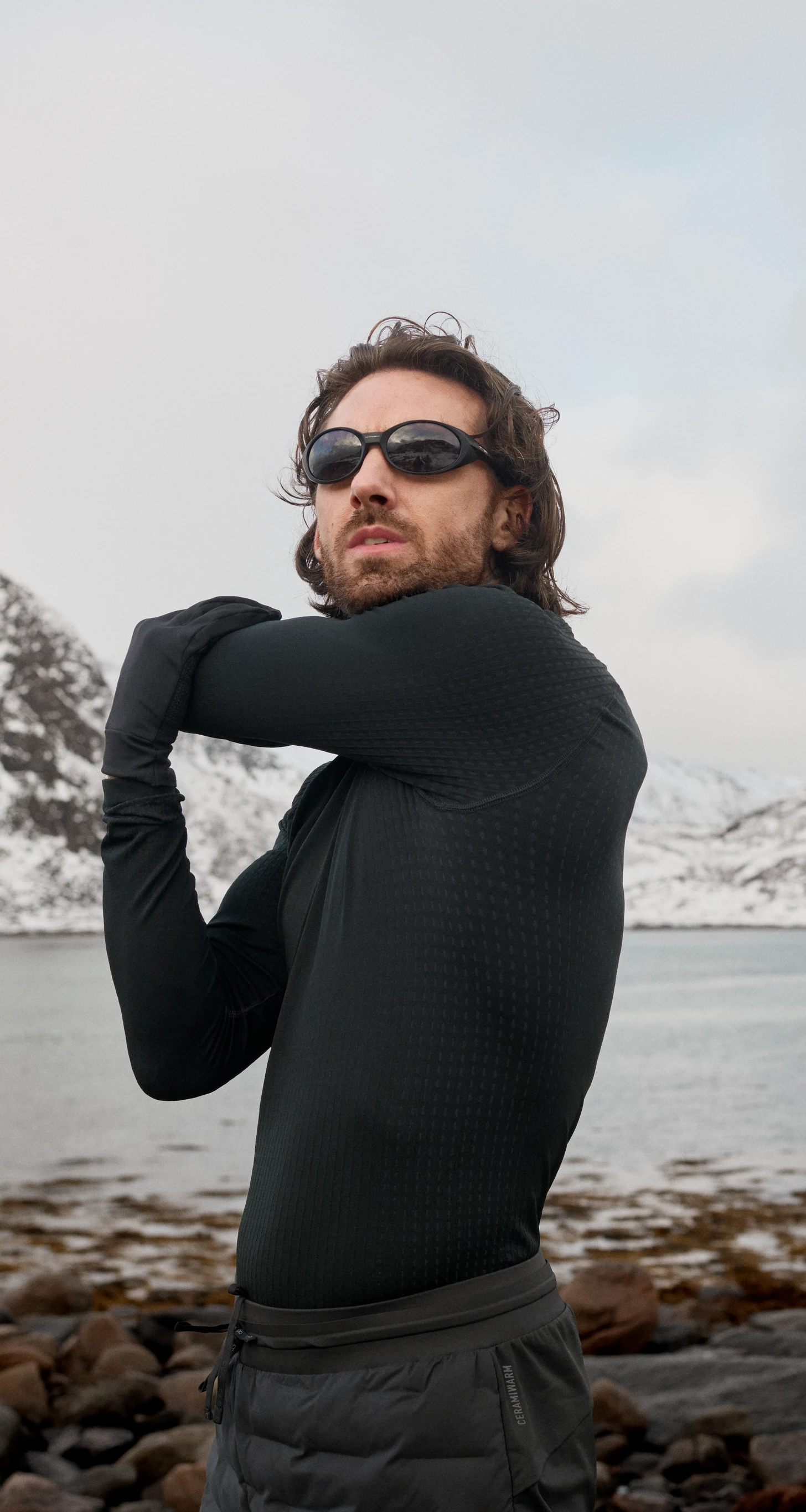 Un uomo con la barba, in abbigliamento da performance nero e occhiali da sole, allunga il braccio in uno stretching su una riva rocciosa, con montagne innevate sullo sfondo.