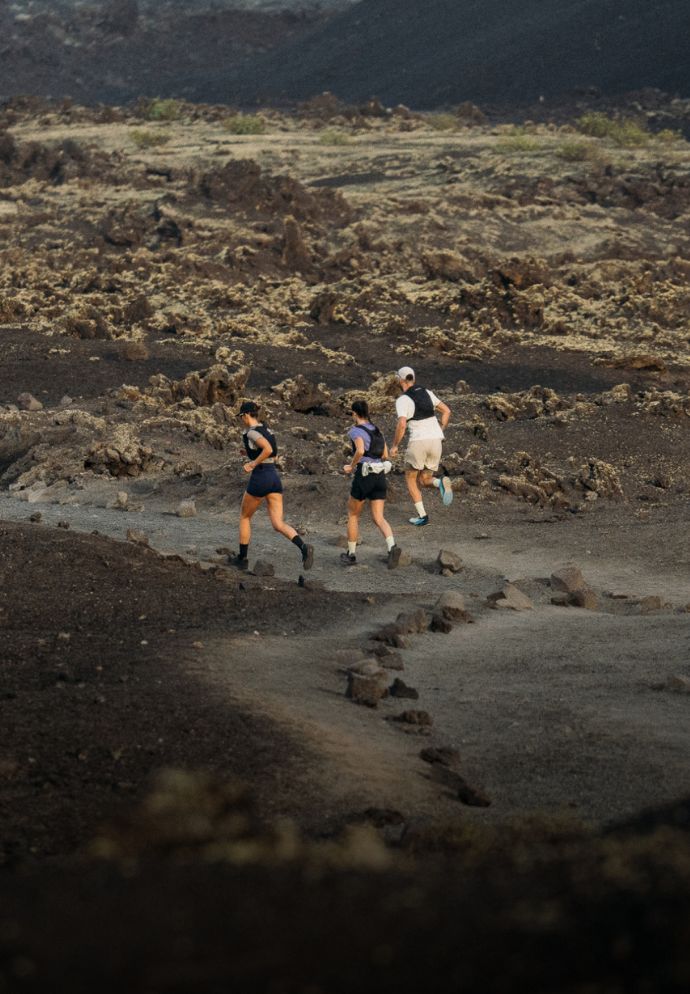 Three people run on a rocky, volcanic trail surrounded by dark brown terrain and scattered stones under a cloudy sky.