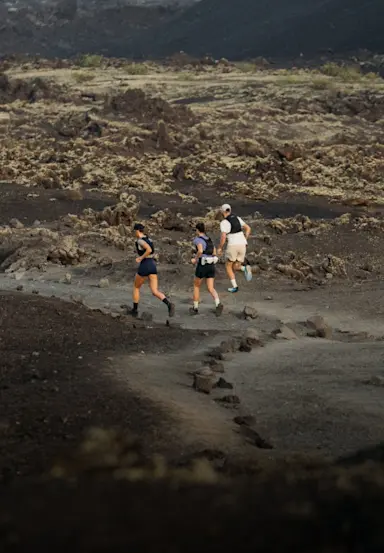 Three people run on a rocky, volcanic trail surrounded by dark brown terrain and scattered stones under a cloudy sky.