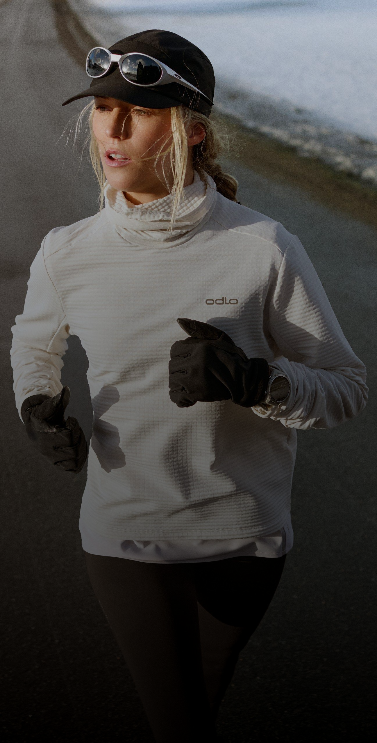 Vrouw rent op een verharde weg met sneeuw langs de kant. Ze draagt een wit Odlo-shirt, een zwarte pet, een zonnebril en zwarte handschoenen.