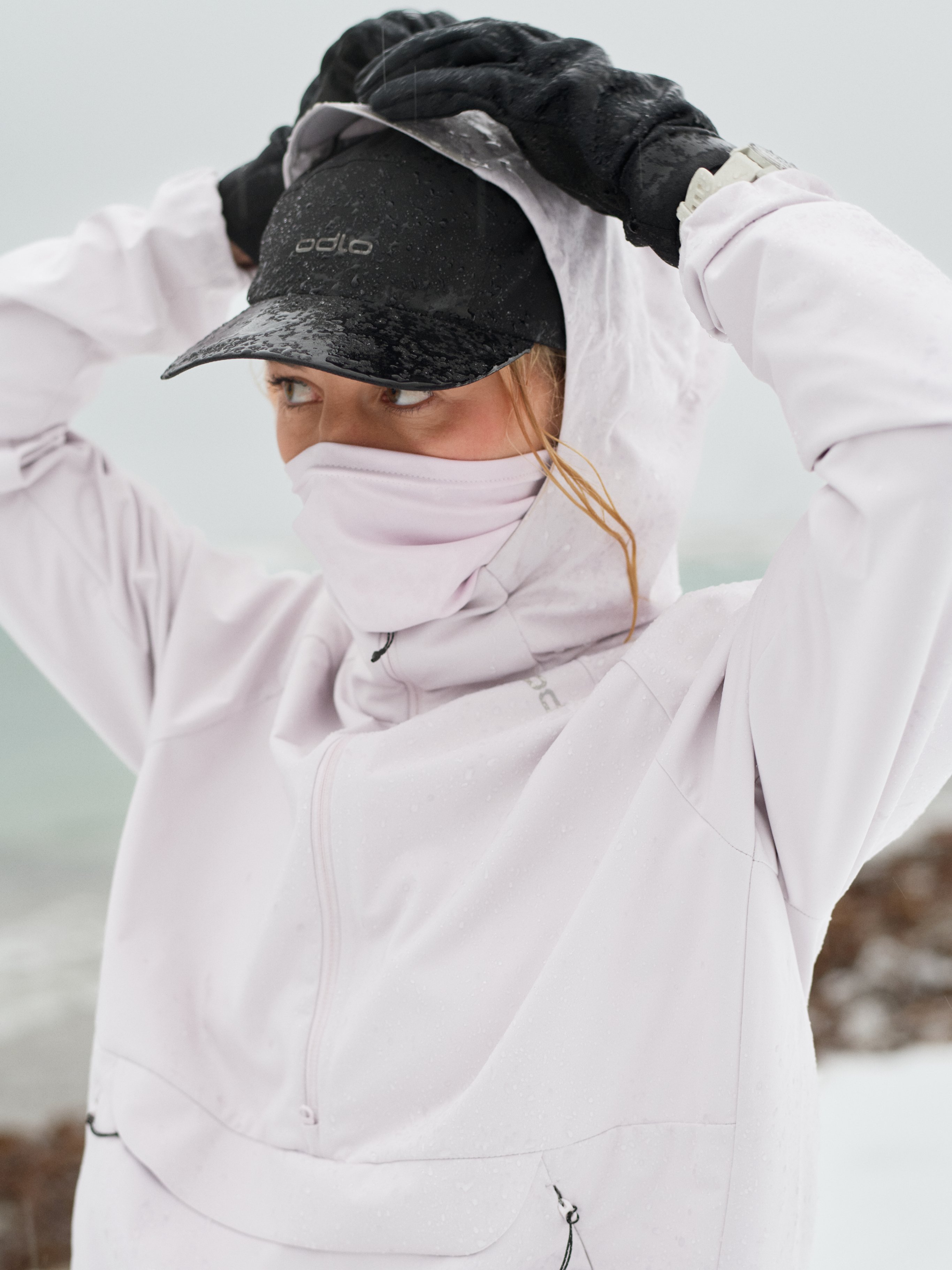 Femme sous la pluie ajustant sa capuche rose sur une casquette noire, portant un cache-cou et des gants noirs.