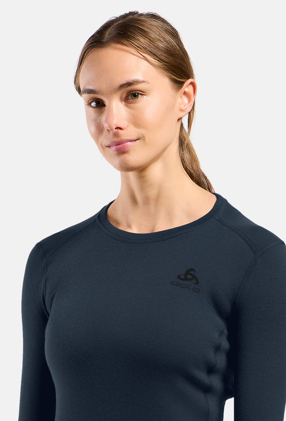 Woman in a navy long-sleeve crew-neck base layer top with small chest logo, posing against a light grey background, hair tied back.