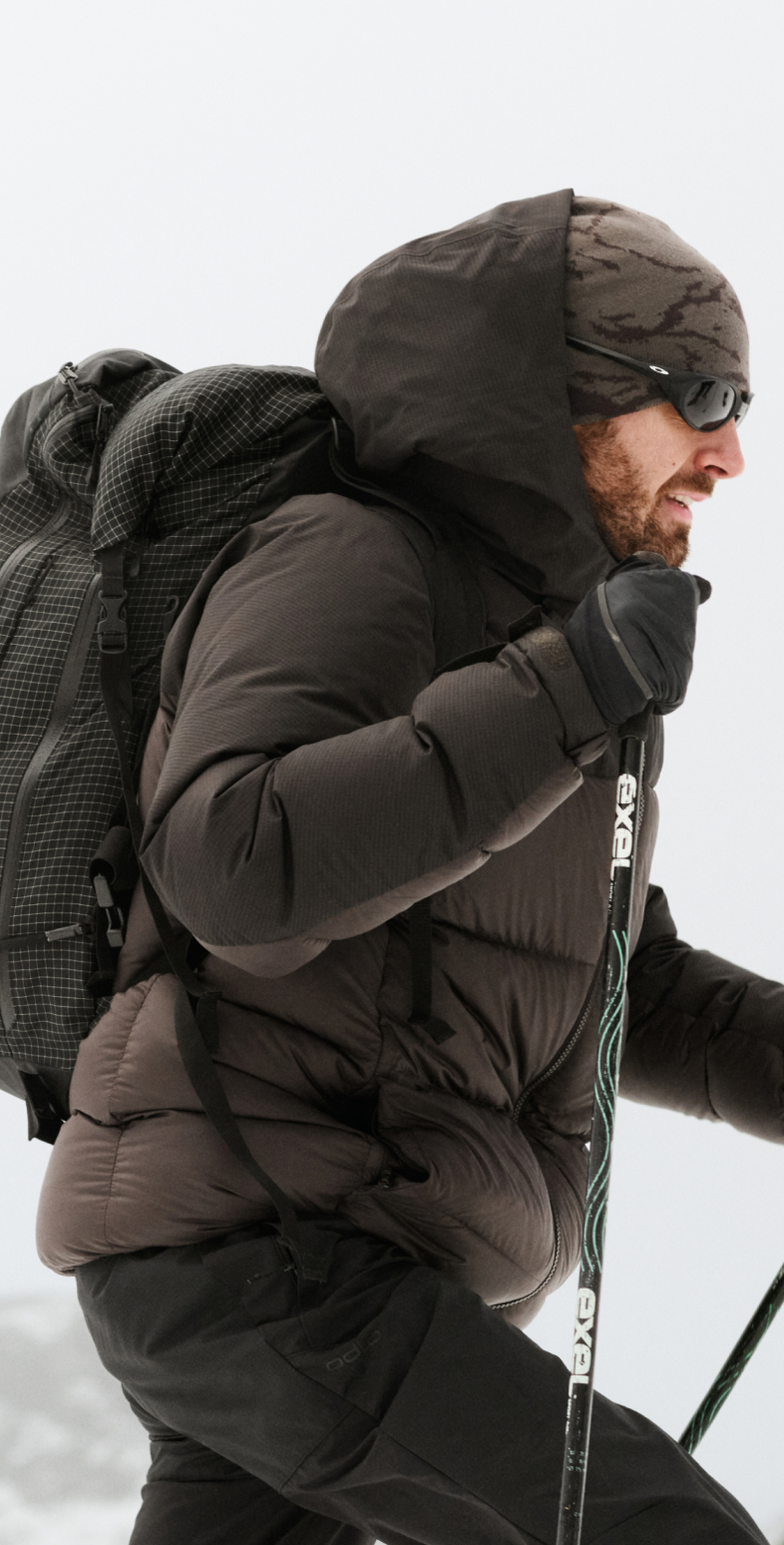 Uomo con la barba che fa trekking nella neve, con indosso un piumino marrone scuro, un berretto mimetico, occhiali da sole, un grande zaino in spalla e bastoncini da trekking.