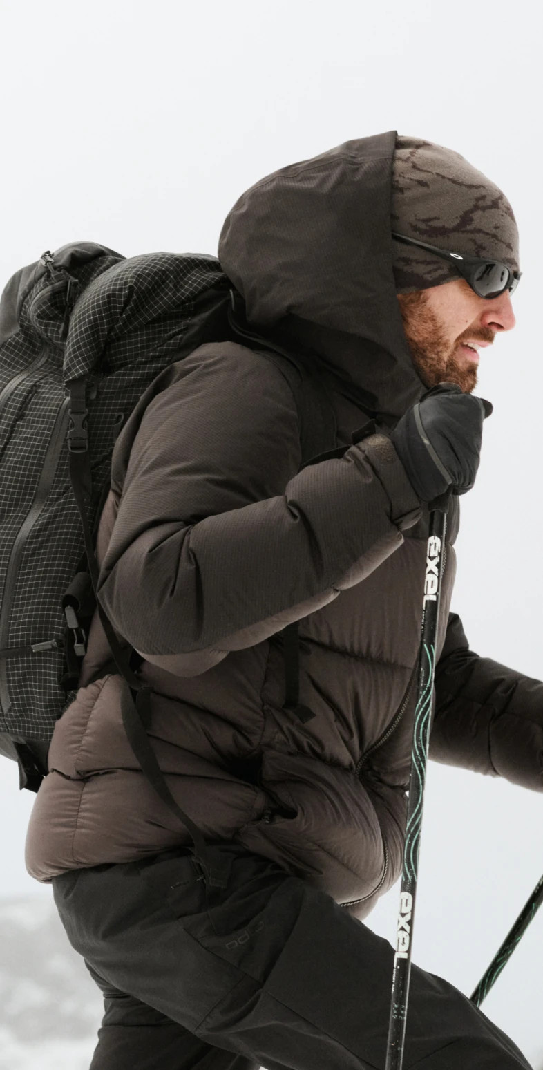 Uomo con la barba che fa trekking nella neve, con indosso un piumino marrone scuro, un berretto mimetico, occhiali da sole, un grande zaino in spalla e bastoncini da trekking.