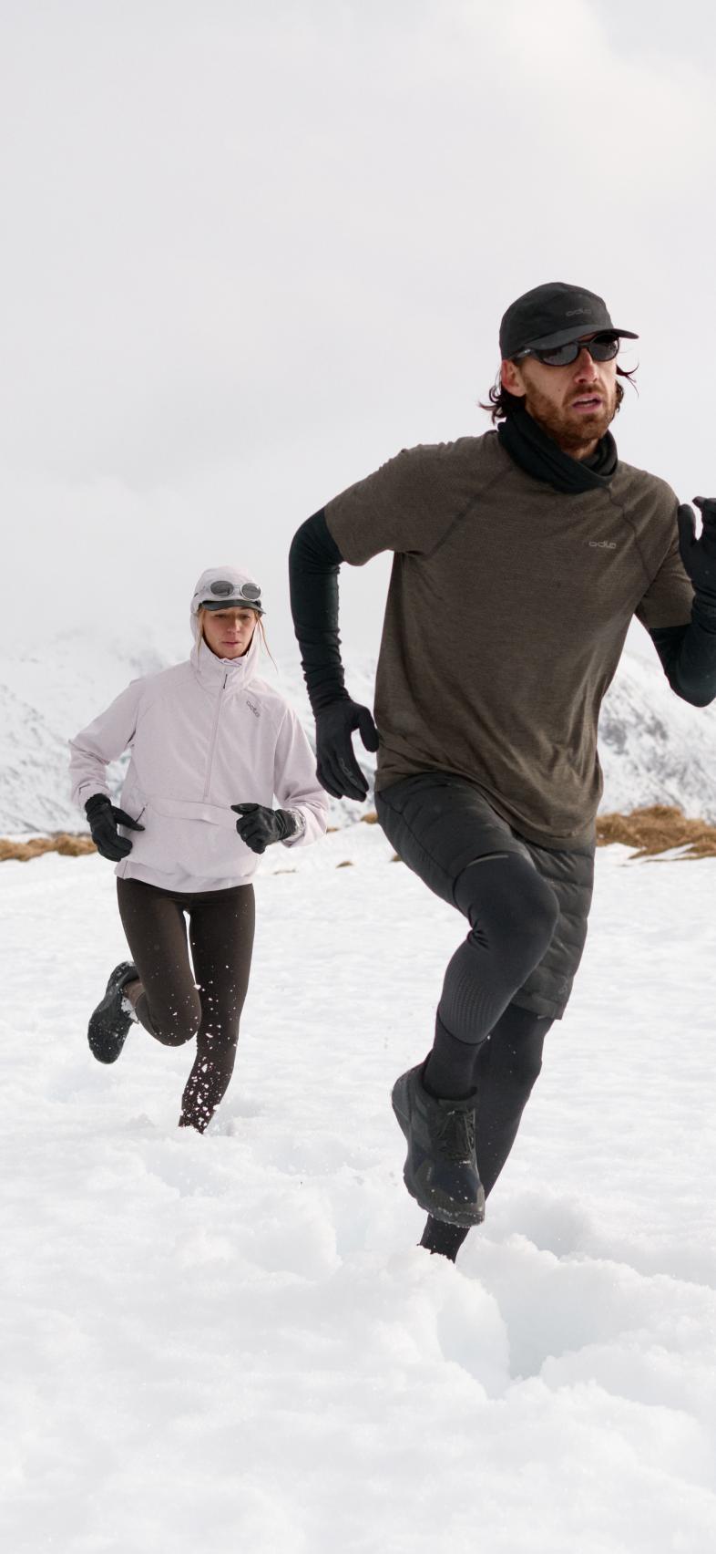 Un homme en chemise marron et une femme en veste blanche courant dans la neige, dans un paysage de montagne hivernal.