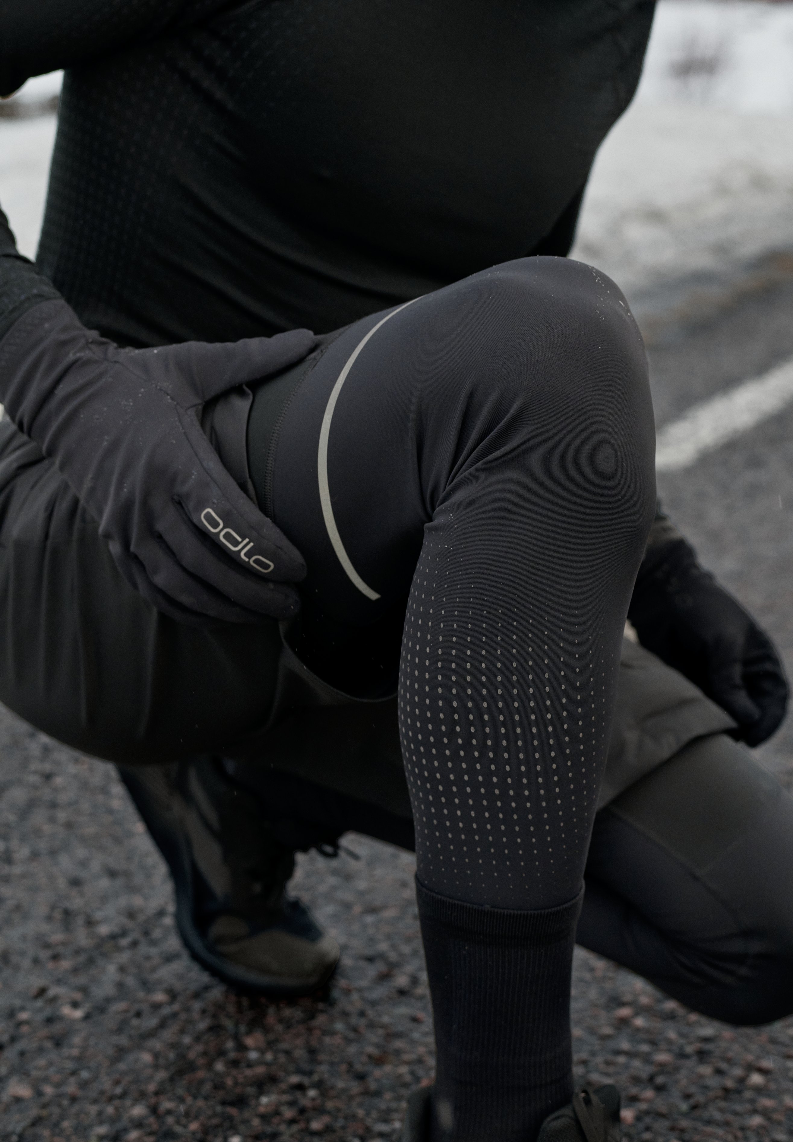 Person kauert in schwarzer Sportbekleidung, trägt Odlo Handschuhe und schwarze Leggings mit reflektierenden Linien und Punkten.